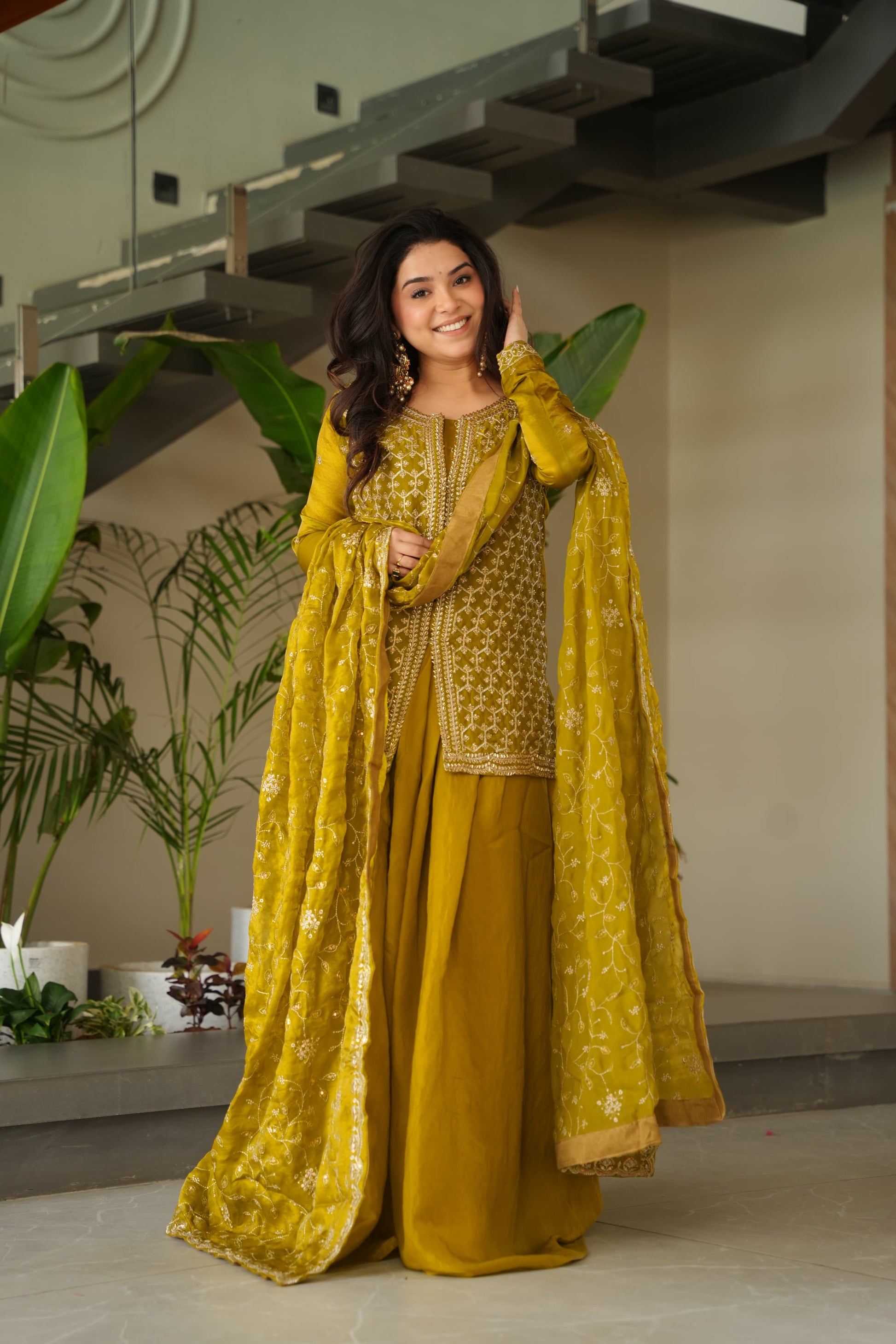 Woman in a yellow traditional outfit standing indoors with plants in the background